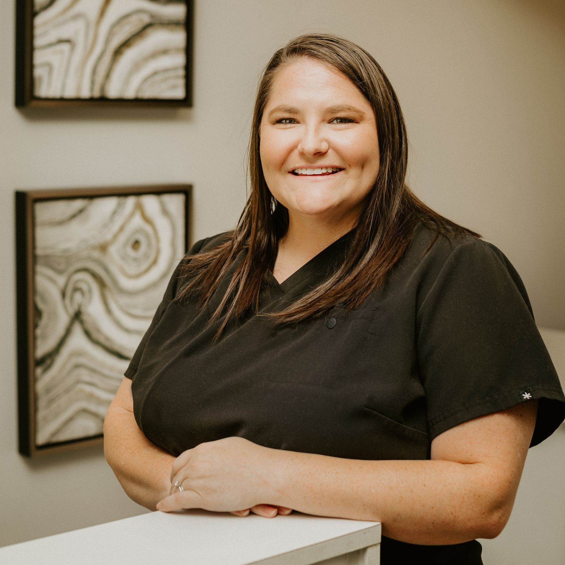 A woman in a black scrub top is smiling while leaning on a counter.