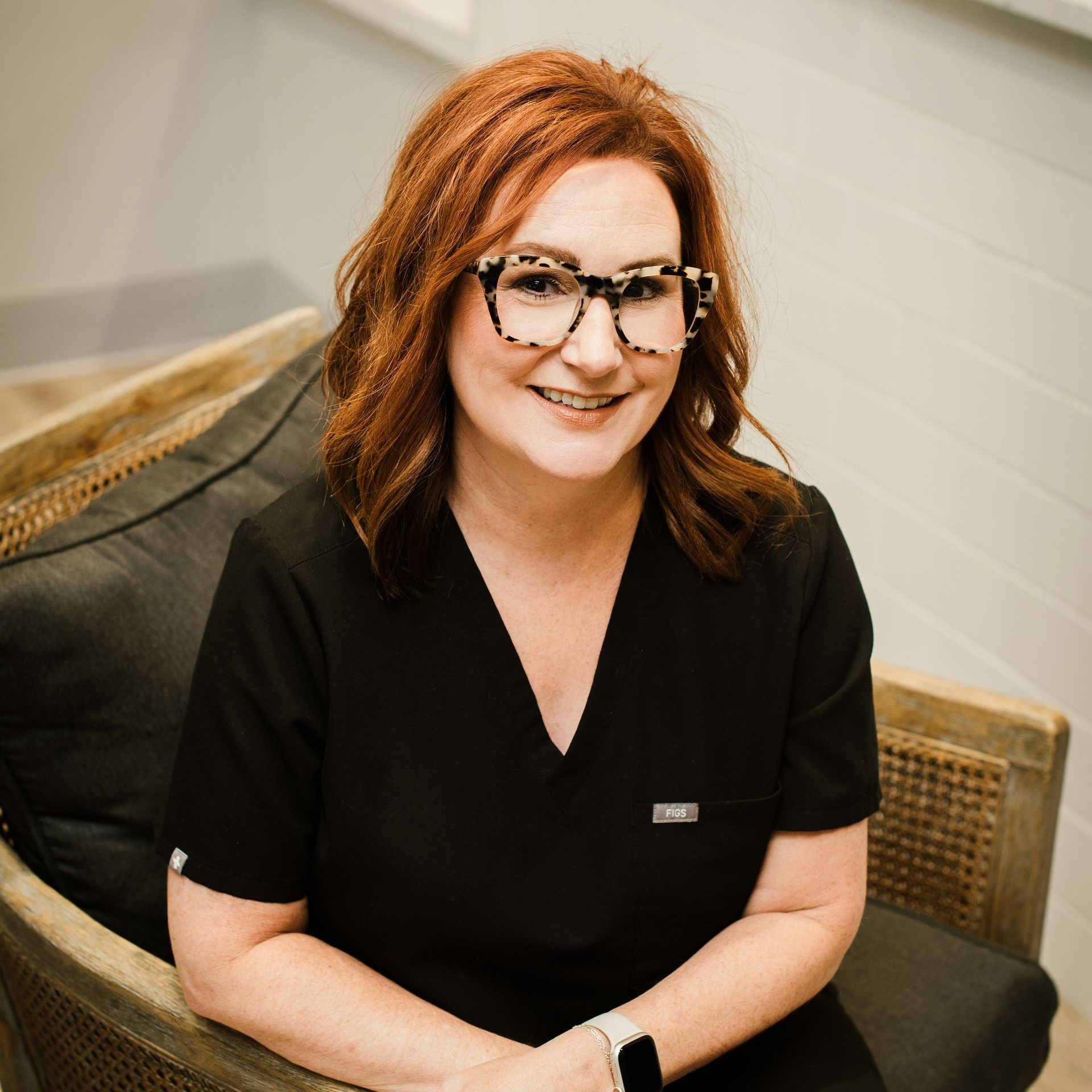 A woman wearing glasses and a black scrub top is sitting in a chair.