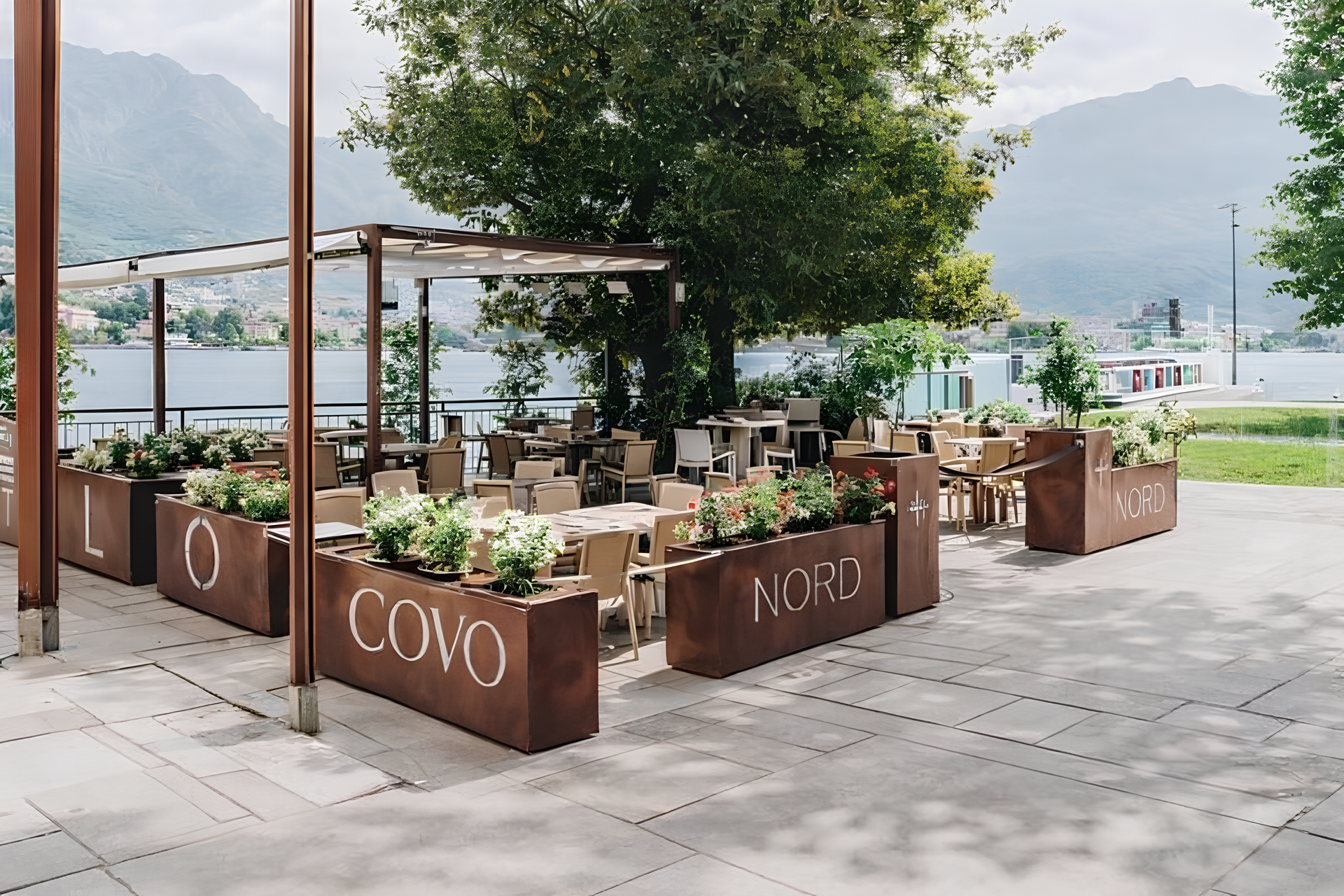 Posti a sedere all'aperto nel ristorante con fioriere in metallo, tavoli e vista sul lago.