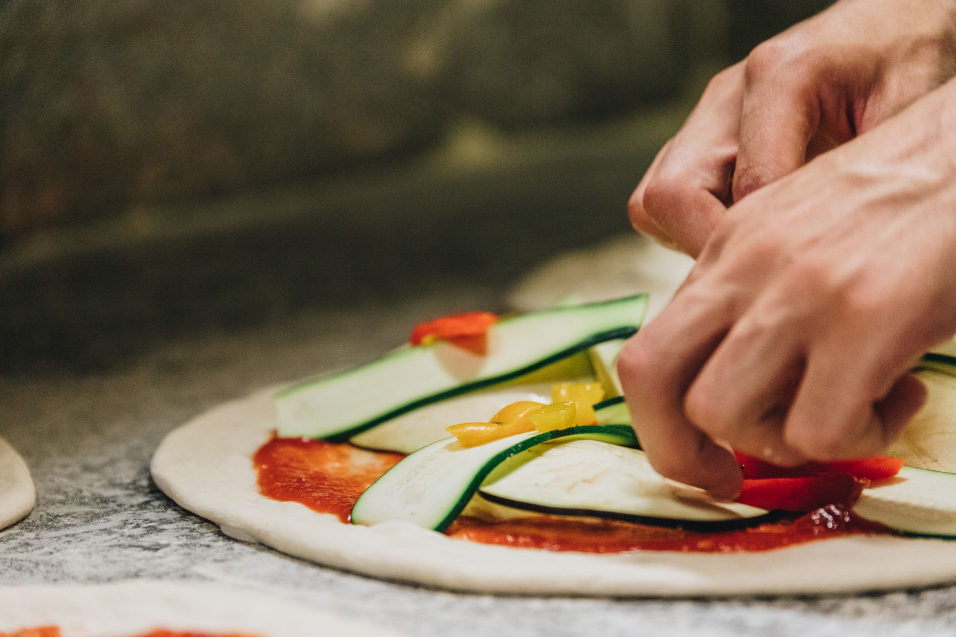 Mani che adagiano sottili fette di zucchine e peperoni su una base per pizza ricoperta di salsa di pomodoro.
