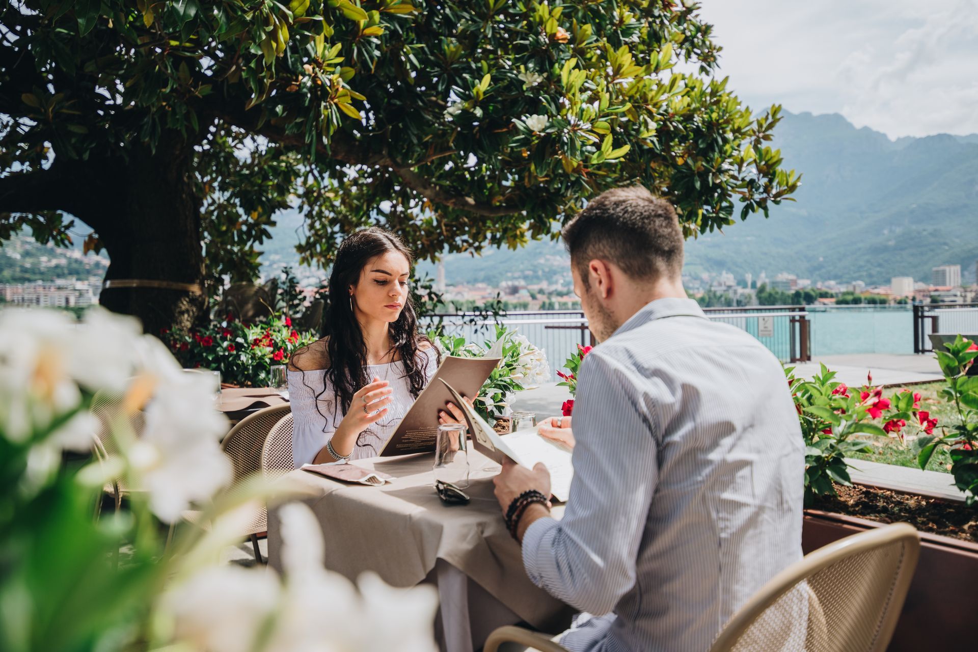 Una coppia consulta i menù seduta a un tavolo all'aperto di un ristorante con vista su un lago e sulle montagne.