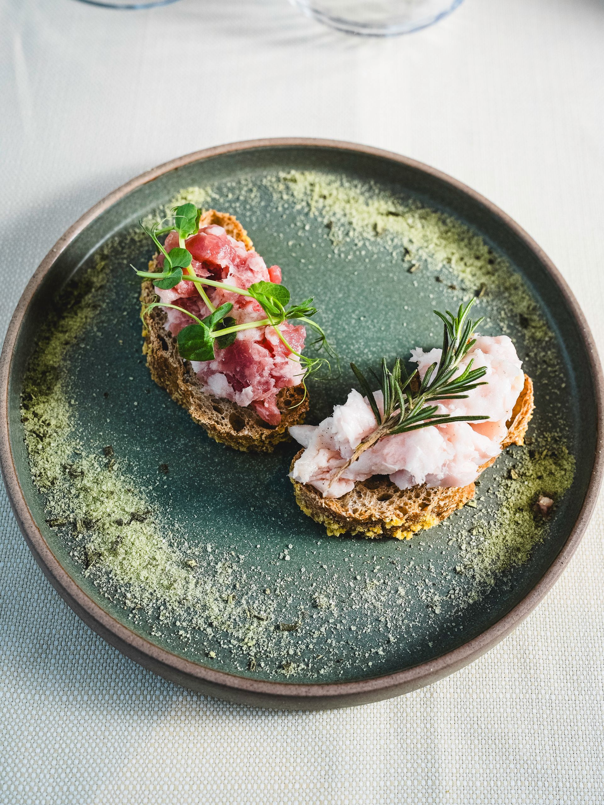 Due crostini guarniti con pesce crudo ed erbe aromatiche fresche sono adagiati su un piatto salvia