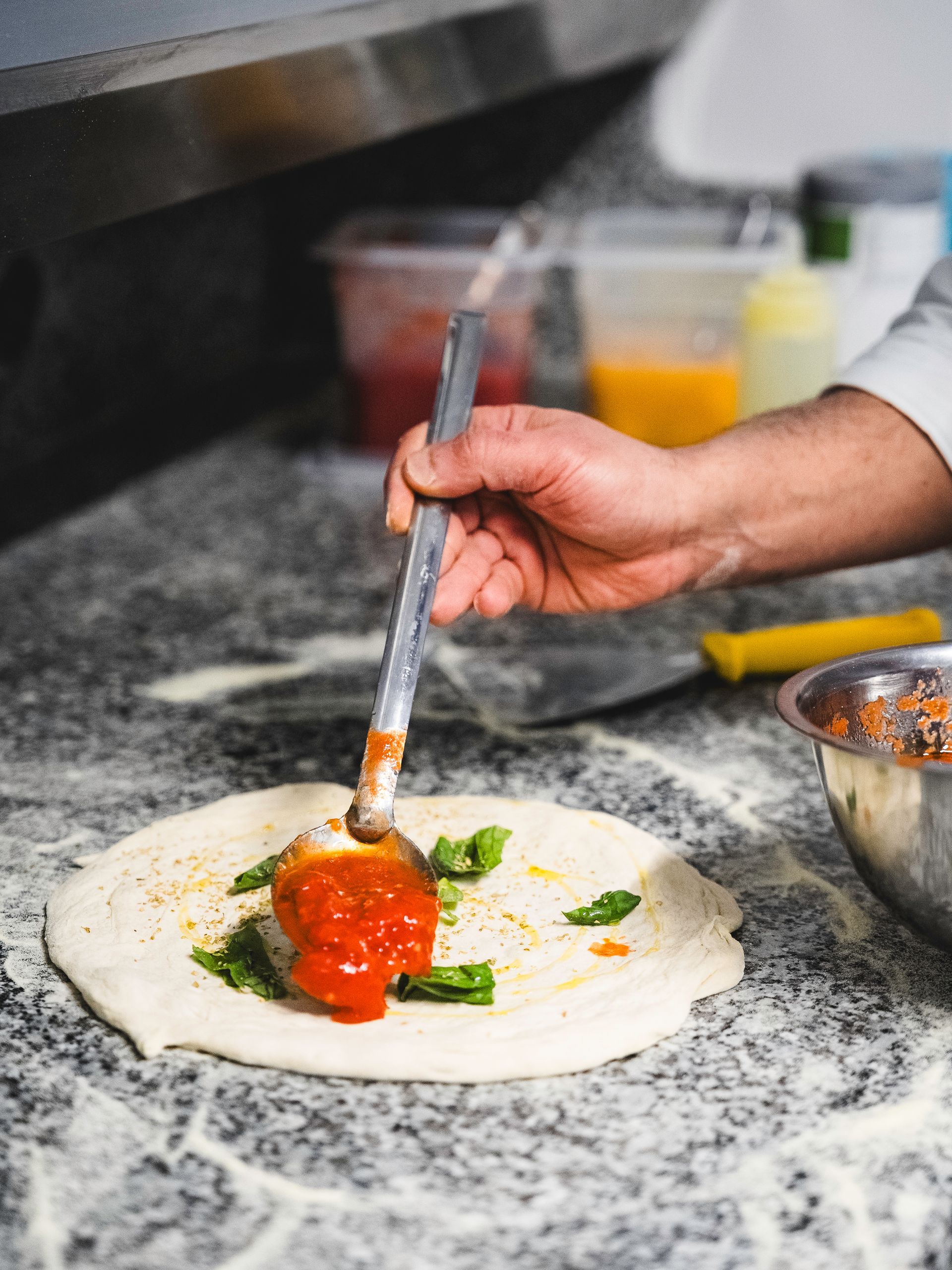 La mano di uno chef usa un mestolo per stendere la salsa di pomodoro sull'impasto della pizza
