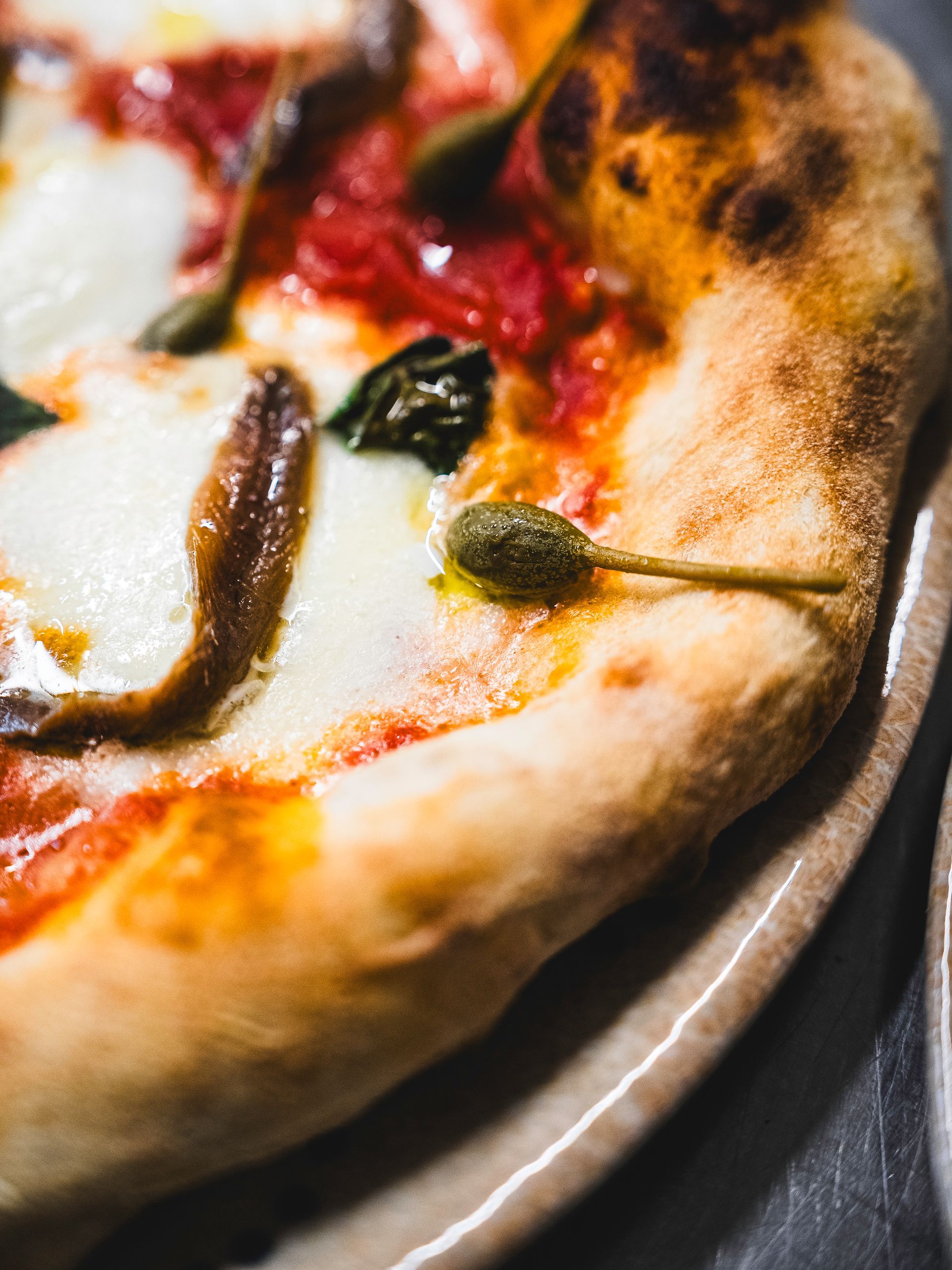 Primo piano di una pizza su un piatto. Ha mozzarella, salsa, acciughe e capperi su una crosta dorata.