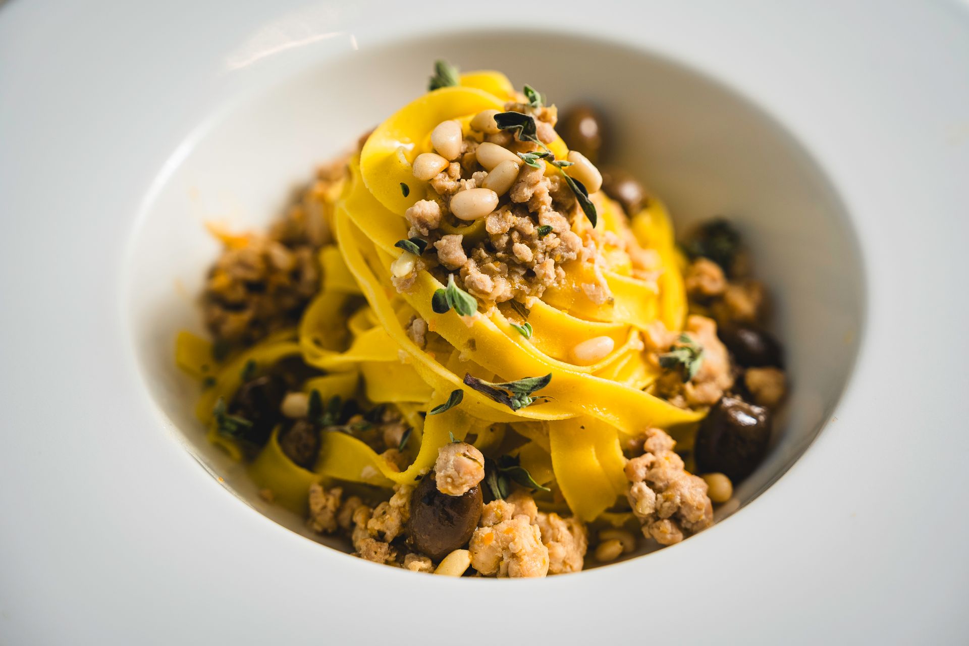 Piatto di pasta con tagliatelle gialle, carne, olive, pinoli ed erbe aromatiche in una ciotola bianca.