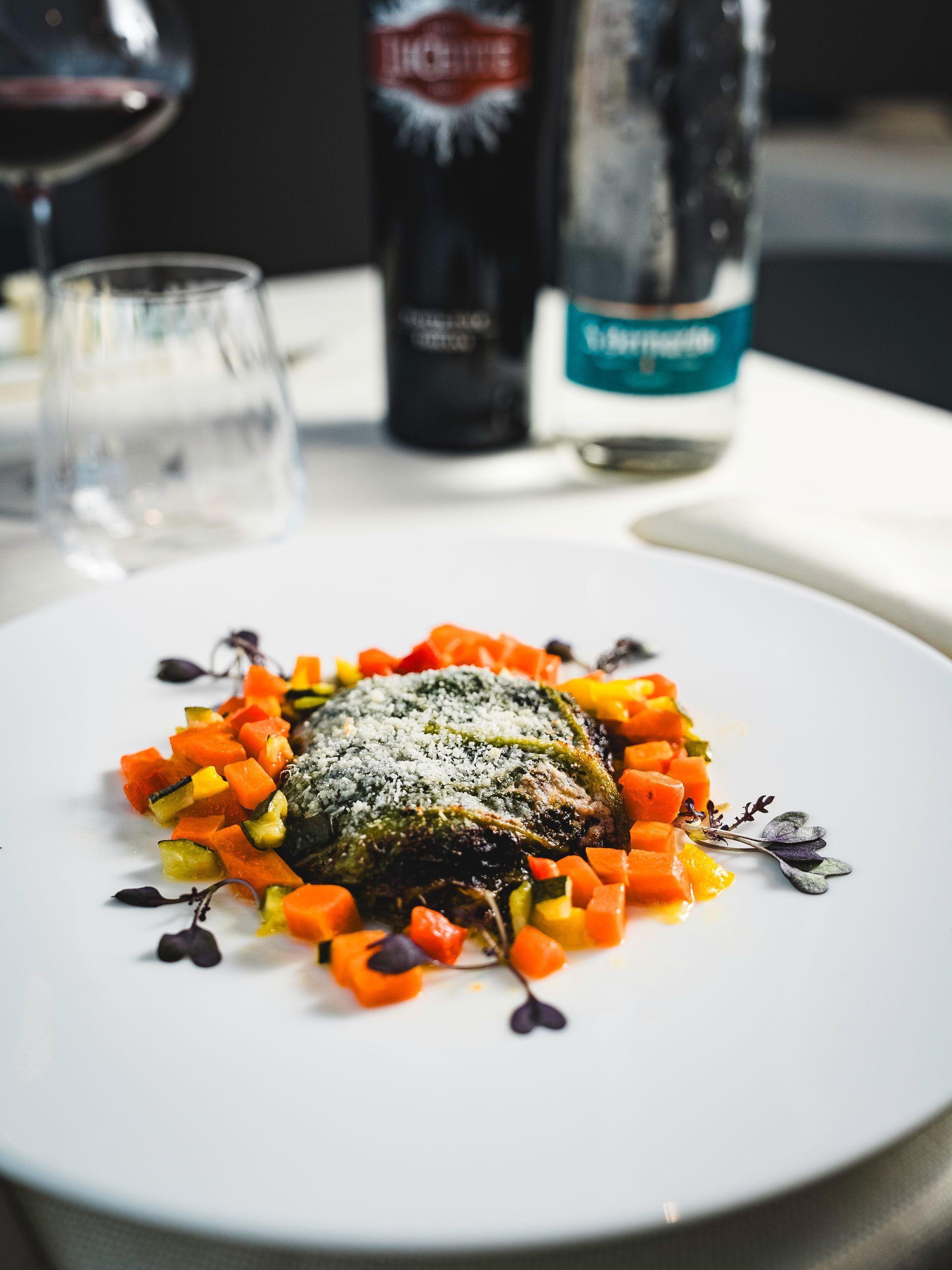 Piatto di cibo guarnito con formaggio, circondato da verdure a cubetti. Bicchieri di vino e bottiglie sullo sfondo.