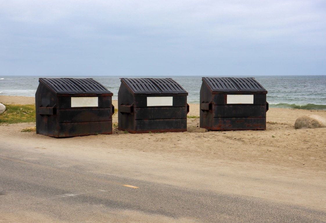 Three black dumpsters on a beach with the ocean in the background under a cloudy sky.