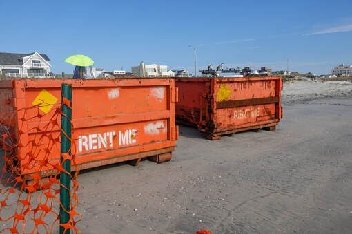 Two orange dumpsters on a sandy beach, with a building background. One dumpster says 