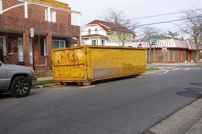 Yellow dumpster on a city street, near buildings and a parked silver truck.