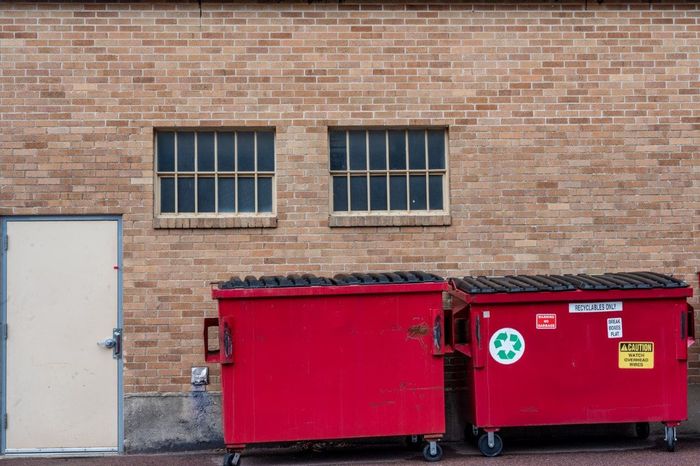Red dumpsters in front of a brick building with windows and a door.