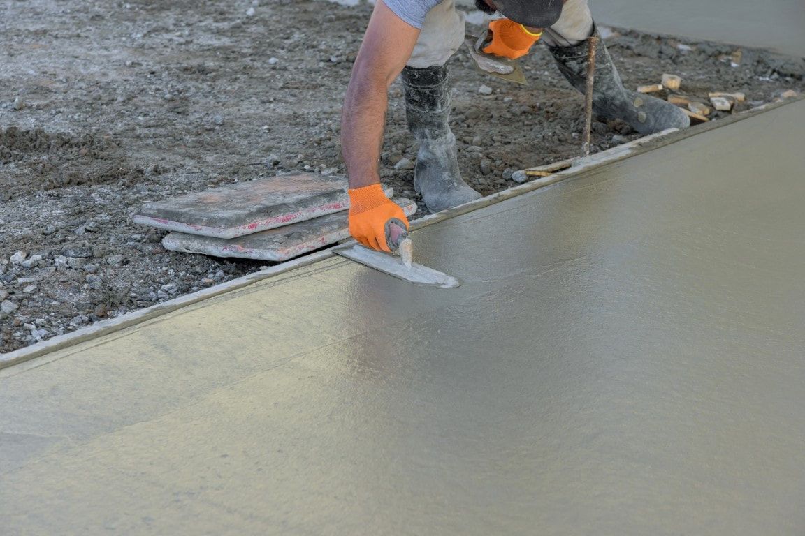Person smoothing wet concrete with a trowel, wearing gloves and boots, on a construction site.