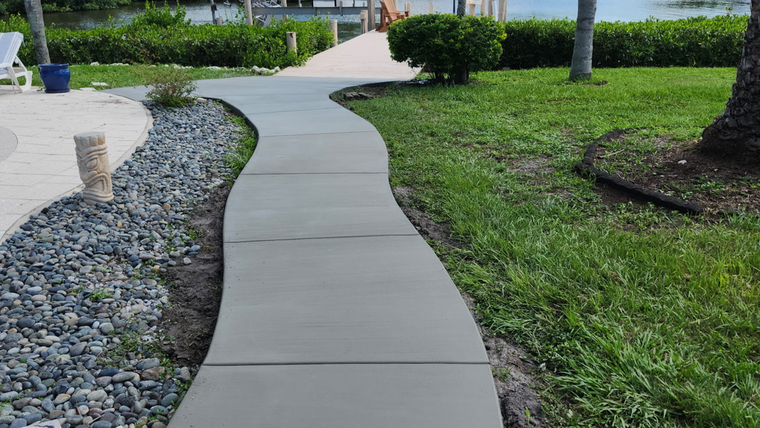 Concrete walkway curves through a grassy lawn toward a dock and water, surrounded by landscaping.