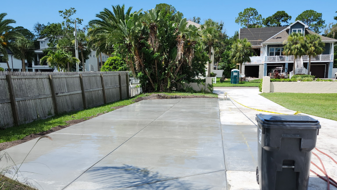 Driveway with gray concrete, a trash can, and houses with palm trees in the background.