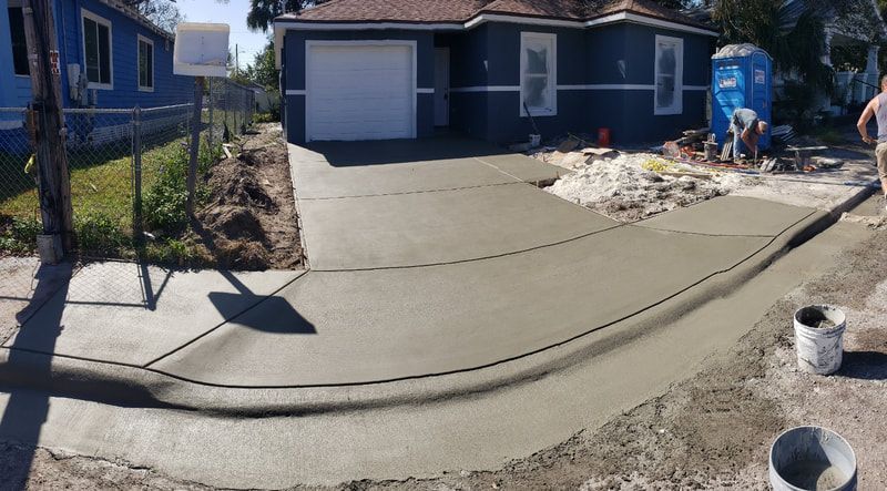 New concrete driveway being poured next to blue house; workers in background.