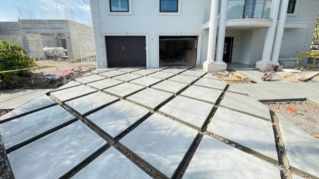 Driveway with large, square concrete tiles with dark gaps, leading to a house with a garage.