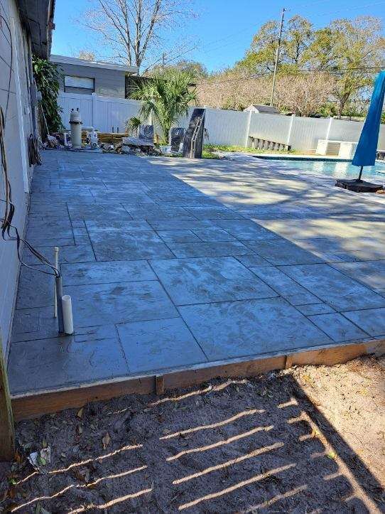 Concrete patio next to a pool under construction, framed by wood, against a white building and fence.