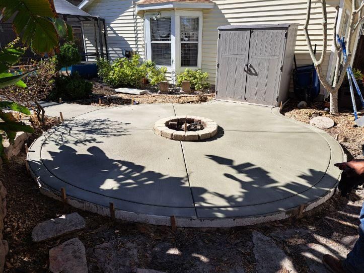 Circular concrete patio with fire pit, near a house and storage shed.