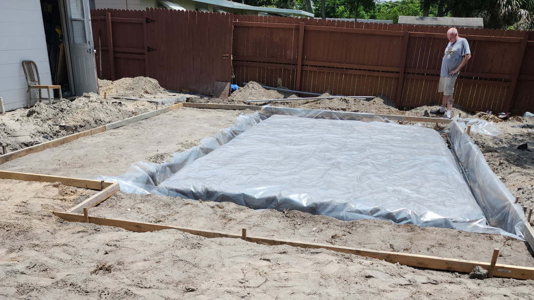 Construction site with person observing. A rectangular area covered with plastic and gravel, surrounded by wooden framing.