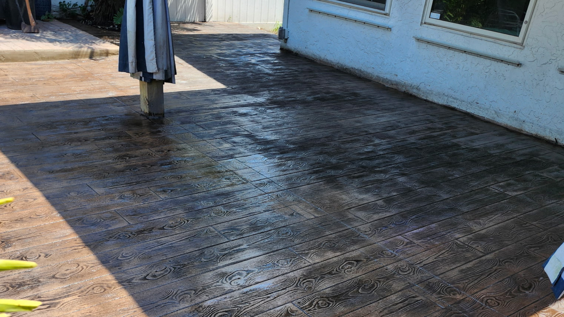 Wet, dark-colored patio with textured pattern next to a white building. Clothesline pole visible.