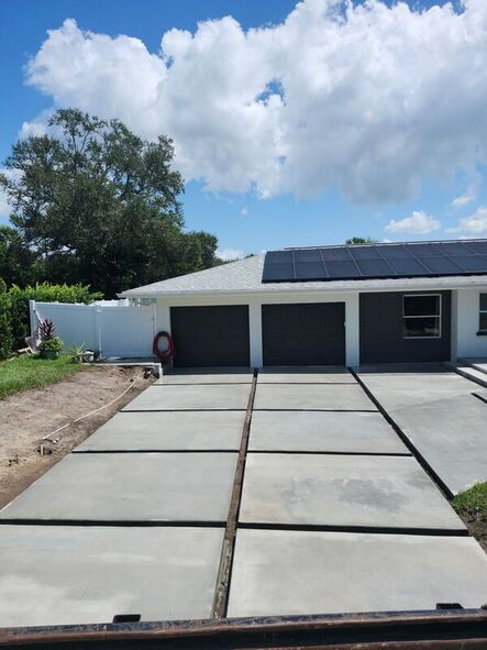 Concrete driveway leading to a white house with two garage doors and solar panels on the roof under a cloudy sky.