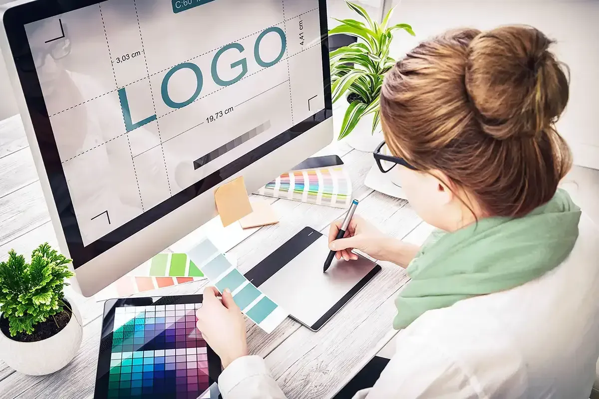 Woman designing a logo on a computer, using a tablet and color swatches at her desk.