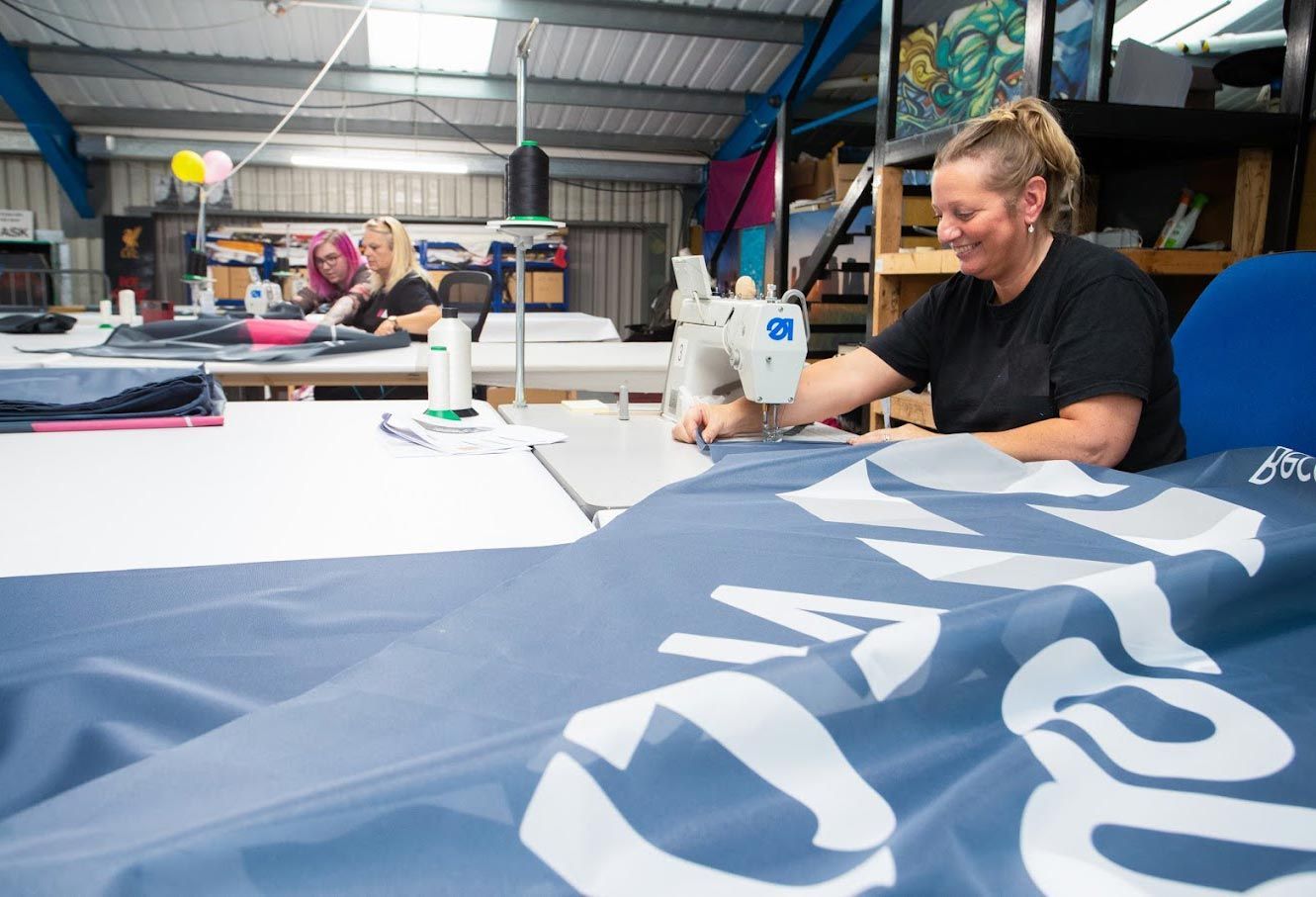 Two women sewing fabric in a workshop. One smiles at a sewing machine, the other works nearby.