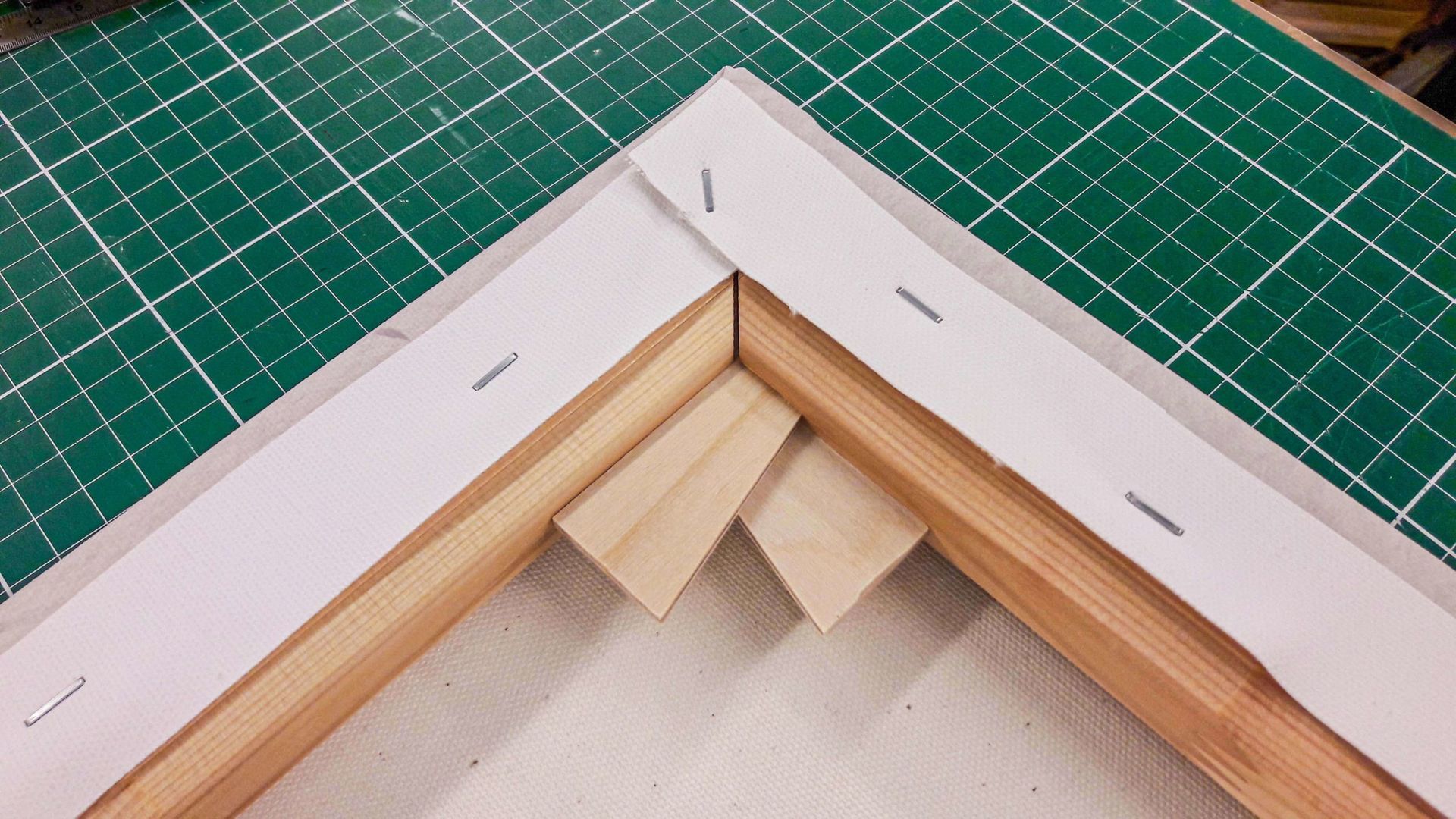 Close-up of a canvas stretched over a wooden frame, showing a corner with staples and wood braces on a green cutting mat.