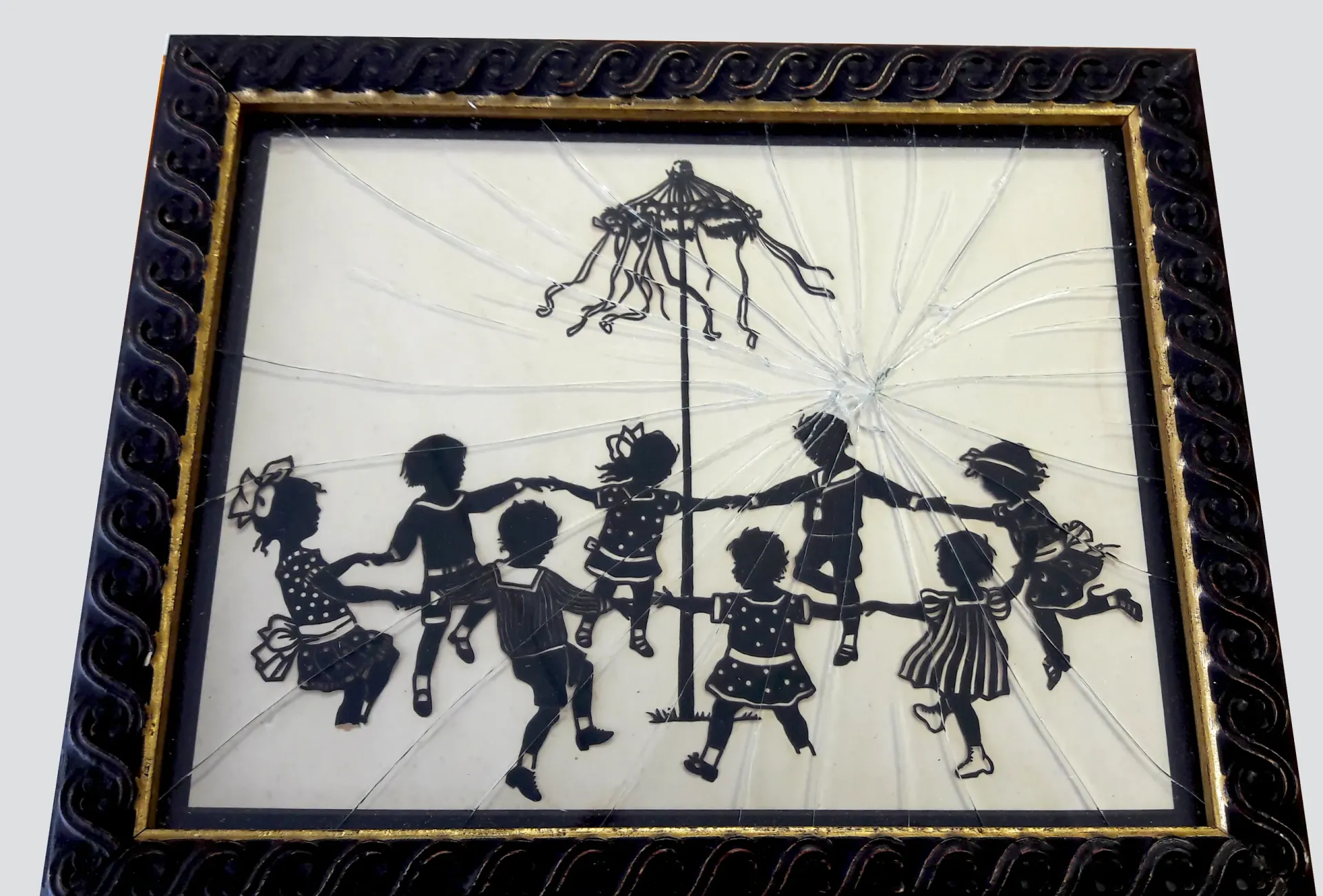 Black silhouette of children dancing around a maypole in a decorative frame.