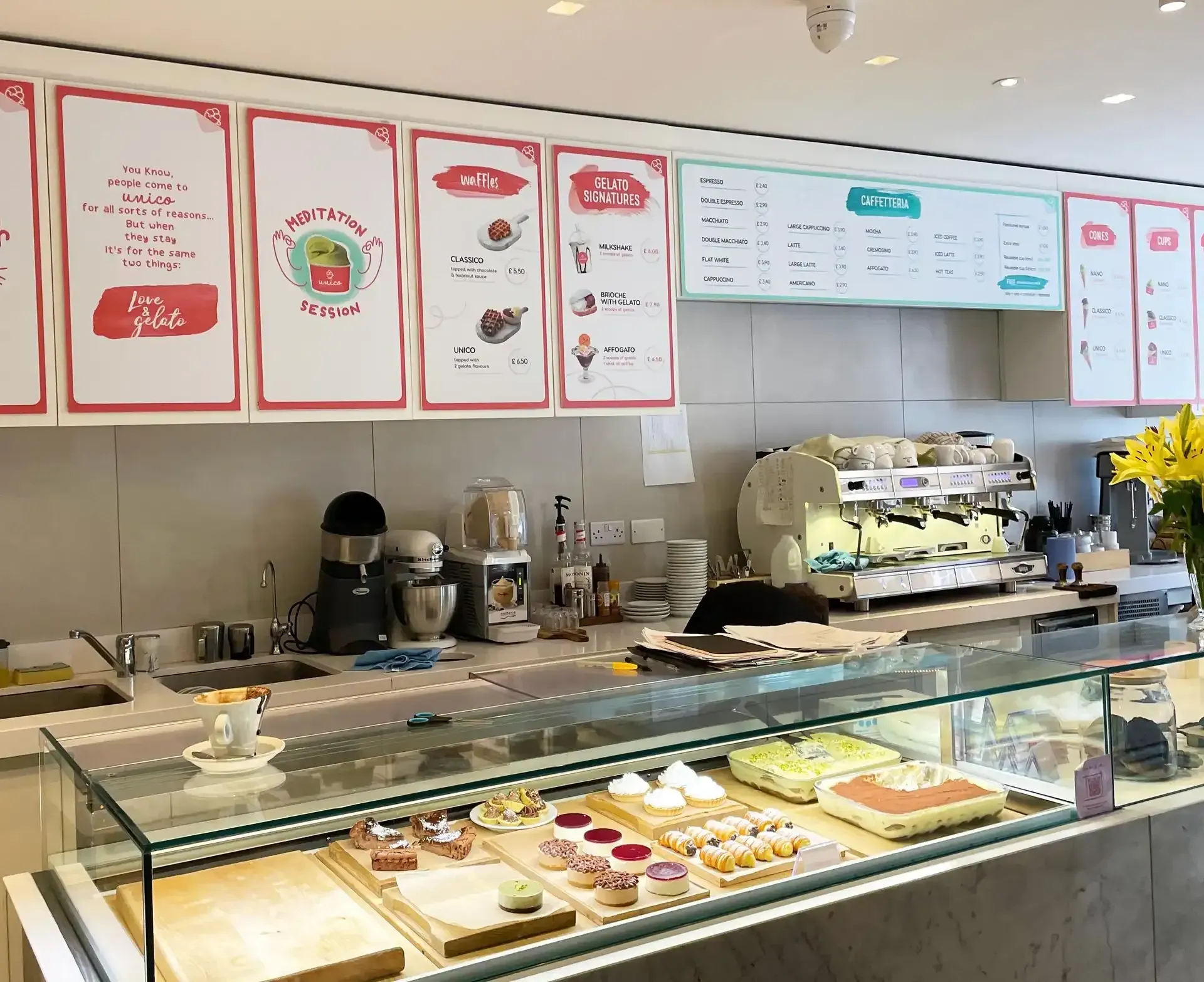 Pastry shop counter with desserts on display, coffee machine, menus, and kitchen equipment.