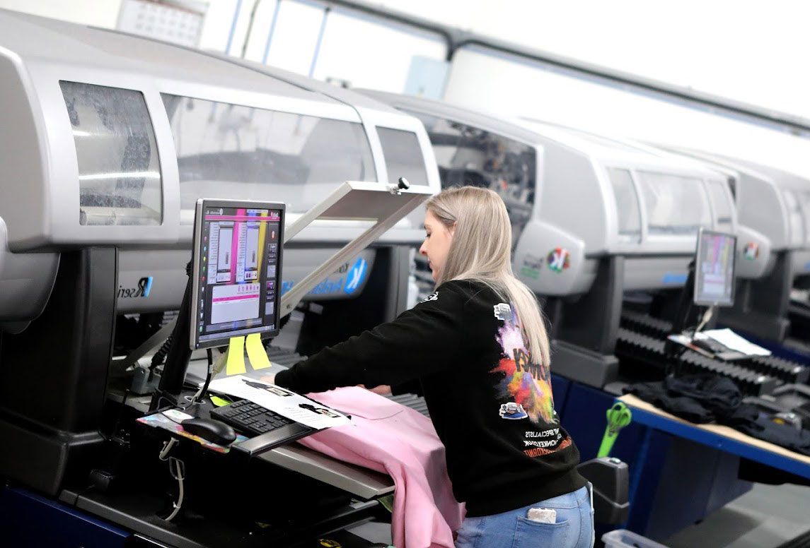Woman operating printing equipment, adjusting a pink garment at a computer. Industrial setting.
