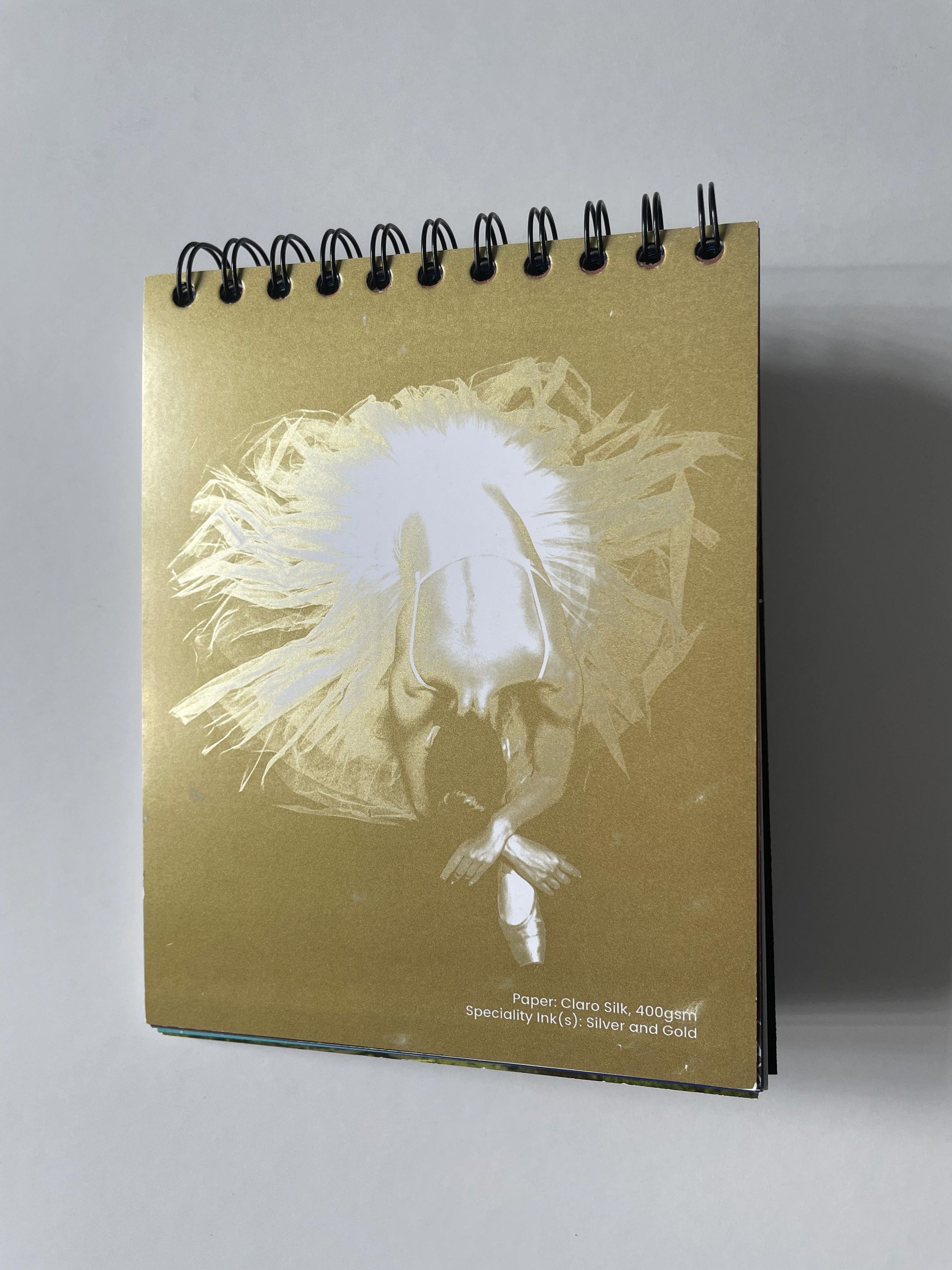 Gold notebook with metallic dandelion design. Spiral bound, leaning on white surface.