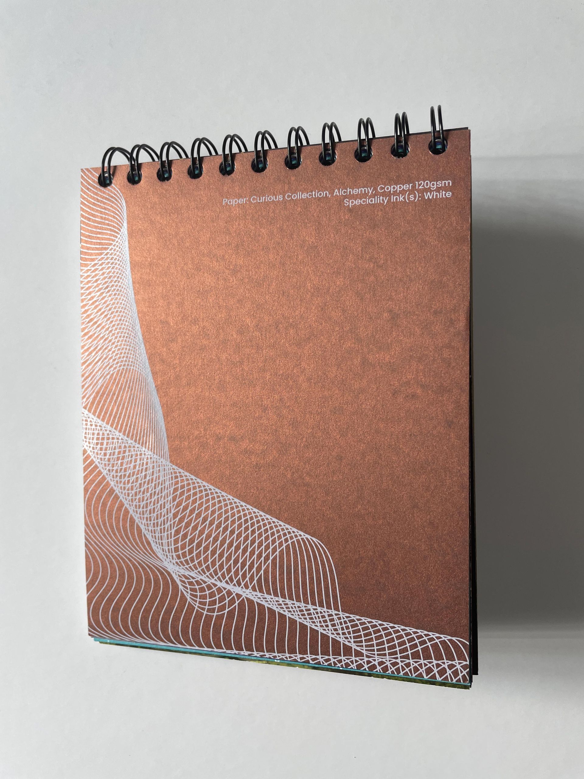 Copper-colored spiral notebook with white abstract lines and text on a white surface.