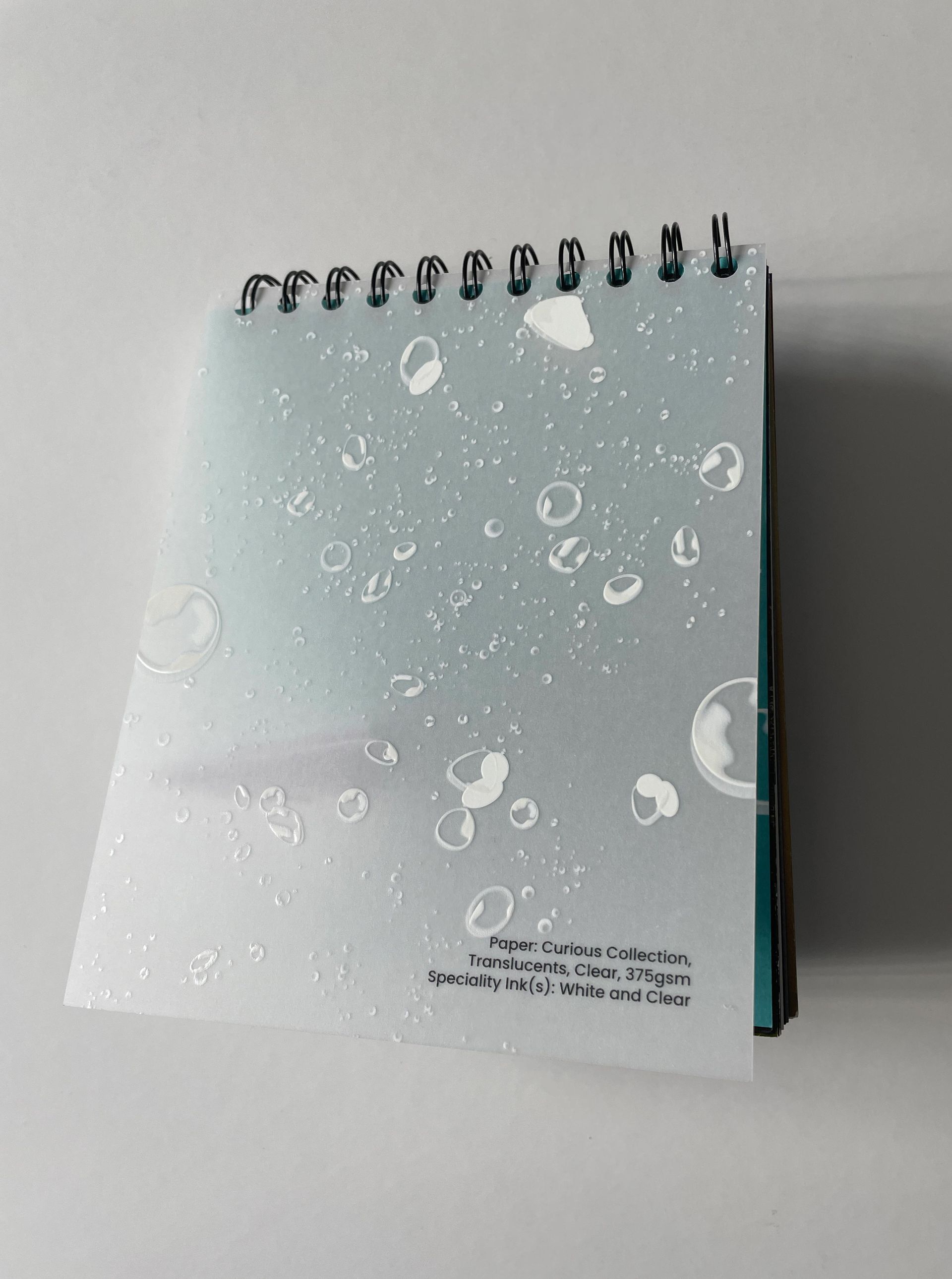 Spiral-bound notepad with water droplet design. Light blue cover with white droplets, resting against a white wall.
