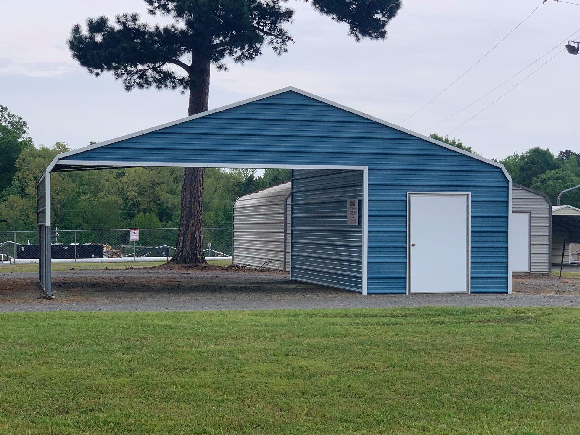 Blue metal carport with a door, under a cloudy sky, on a grassy area.