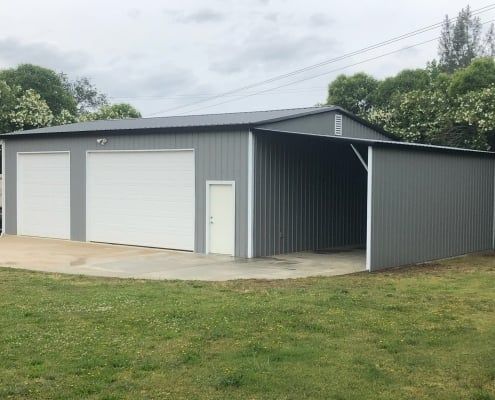 Gray metal garage with two bays and an open-air carport. White doors, concrete pad, and green lawn.