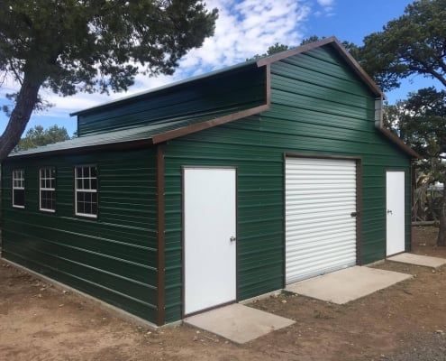 Green metal barn with white doors, windows, and a garage door, under a blue sky.