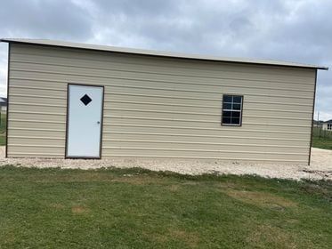 Tan metal storage building with white door and window on gravel base.
