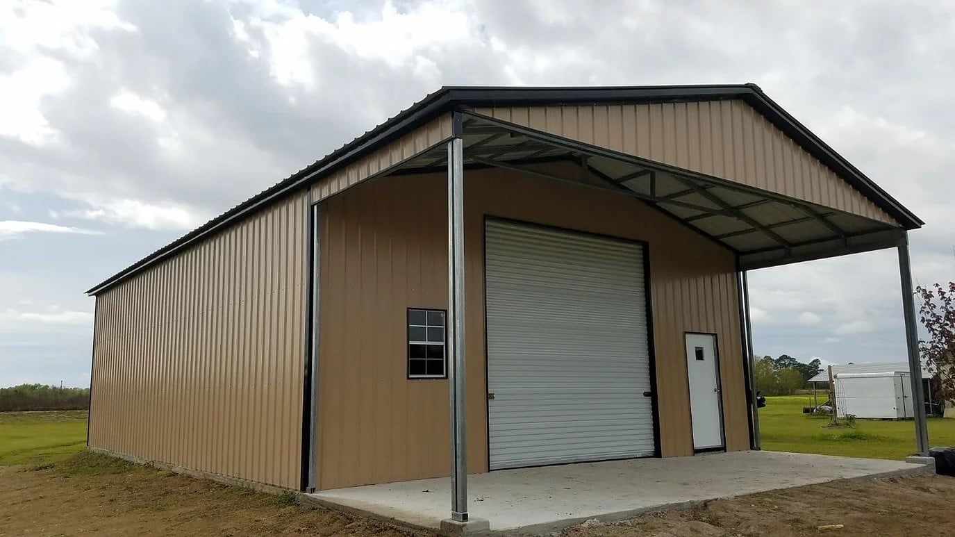 Tan metal barn with a concrete pad, awning, and large roll-up door under a cloudy sky.