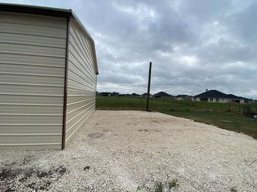 Tan metal shed, gravel area, and a single wooden pole stand against a cloudy sky in a suburban setting.