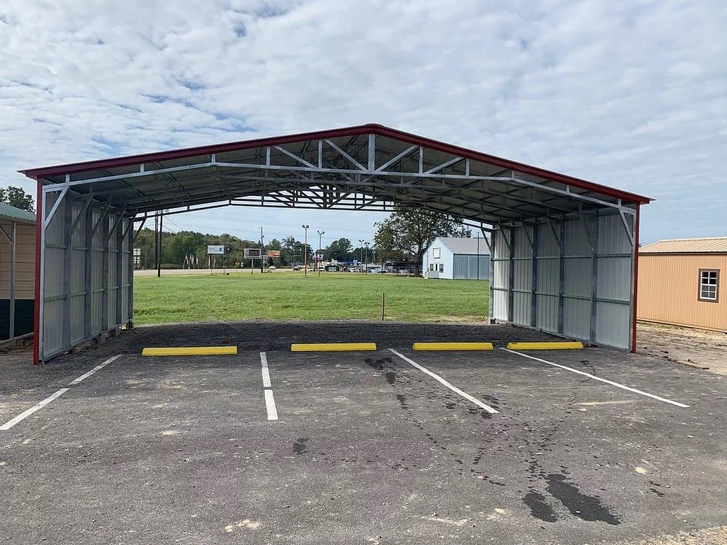 Metal carport with red trim and parking spaces on gravel, overlooking a grassy field.