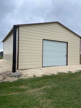 Tan metal garage with brown trim, roll-up door, and gravel base, situated on green grass under a cloudy sky.