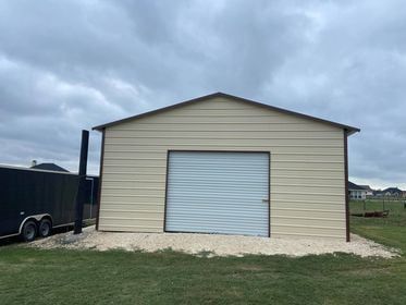 Tan metal shed with a garage door, set on gravel in a grassy yard. Overcast sky.