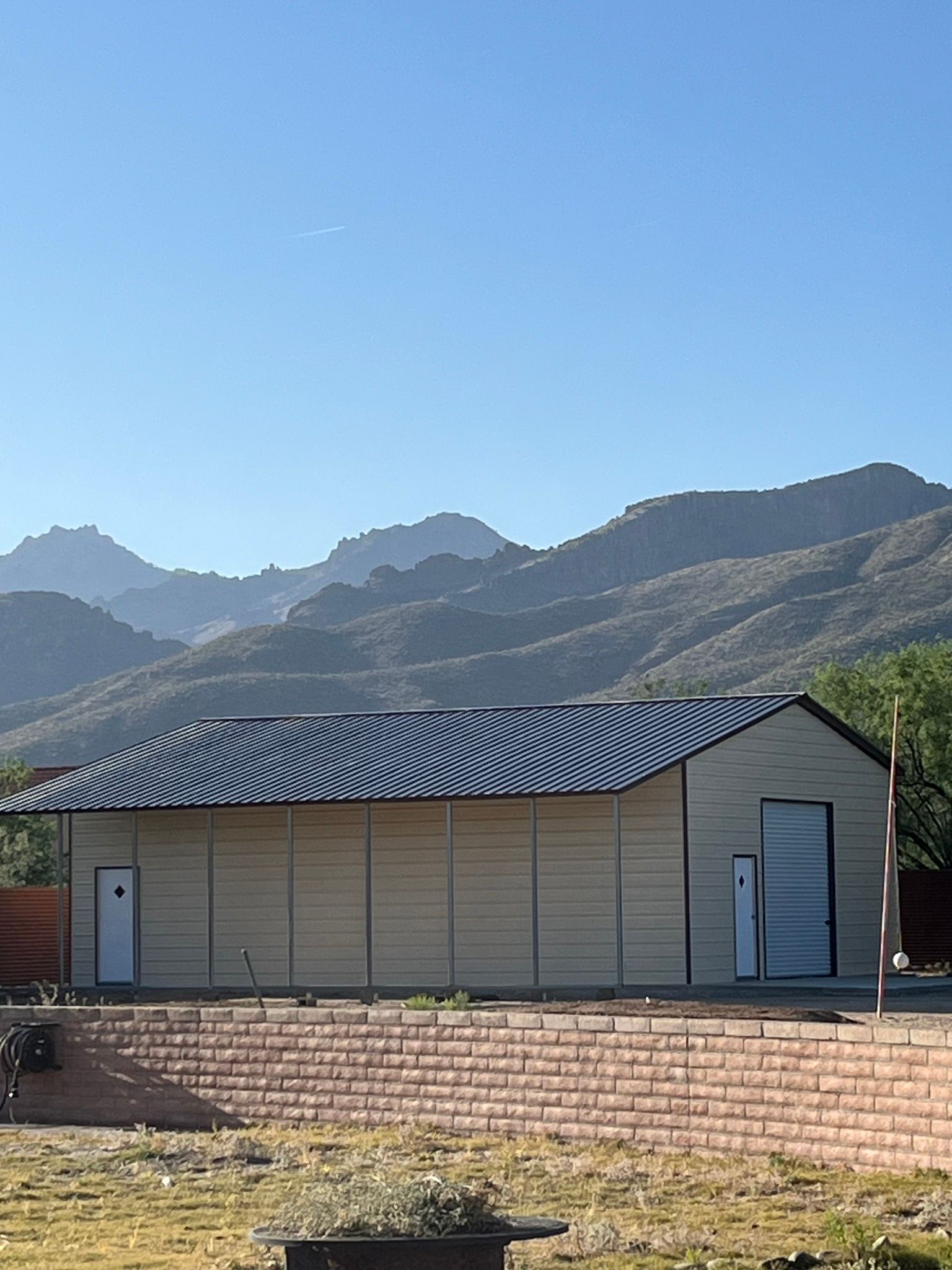 Light-colored building with corrugated roof against a mountain backdrop under a clear sky.