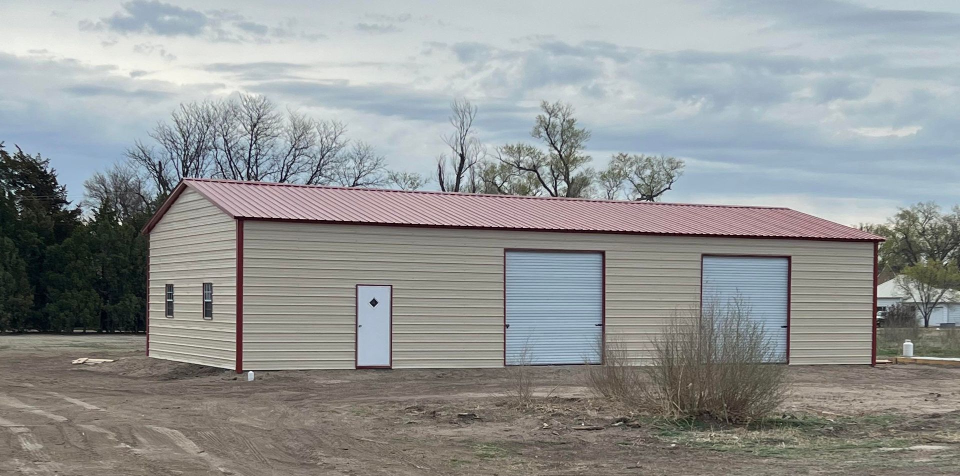 A tan metal building with a red roof, white door, and two garage doors, under a cloudy sky.