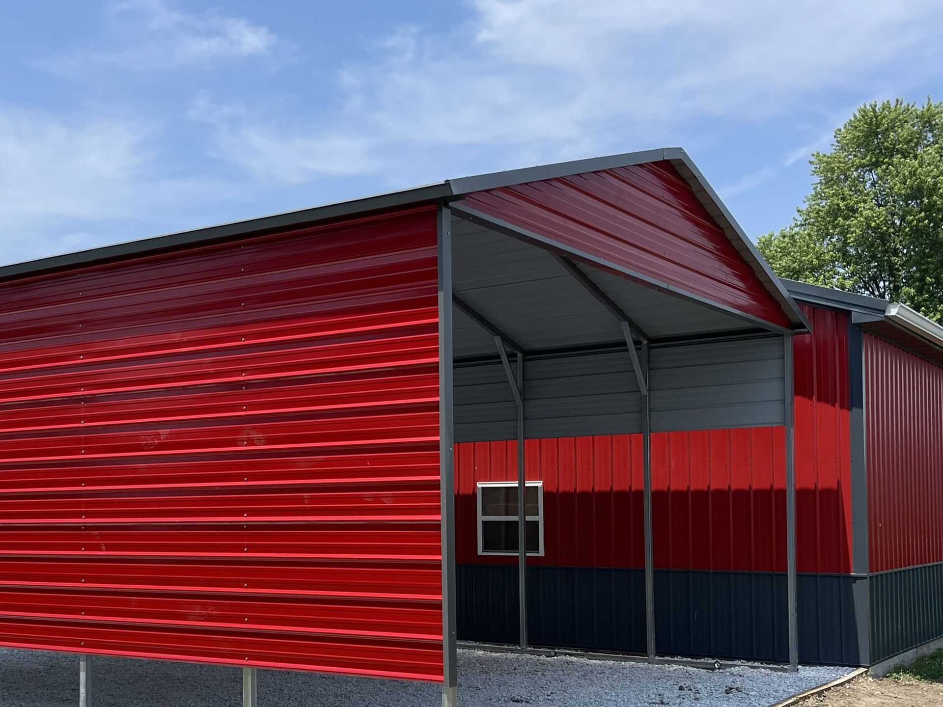 Red and gray metal barn with open side on gravel, blue sky background.