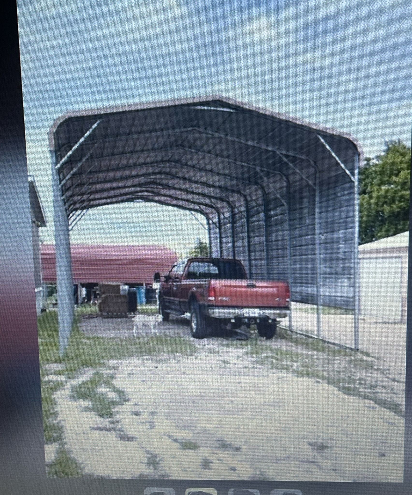 Red pickup truck parked under a large metal carport with a gravel driveway.