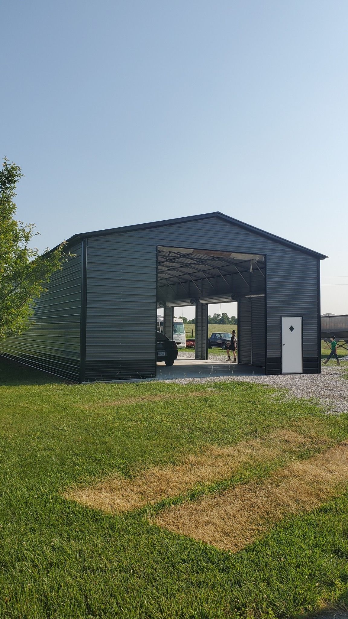 Gray metal shed with open front, white door, and gravel area, on green grass under a clear sky.