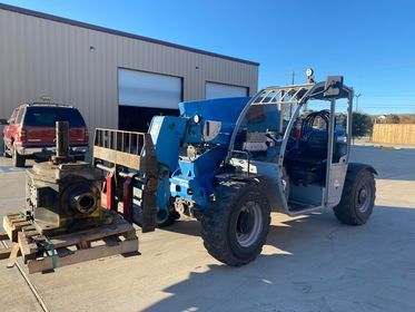 A blue telehandler with a missing engine and parts.  An engine sits on a pallet nearby, outside in a sunny setting.