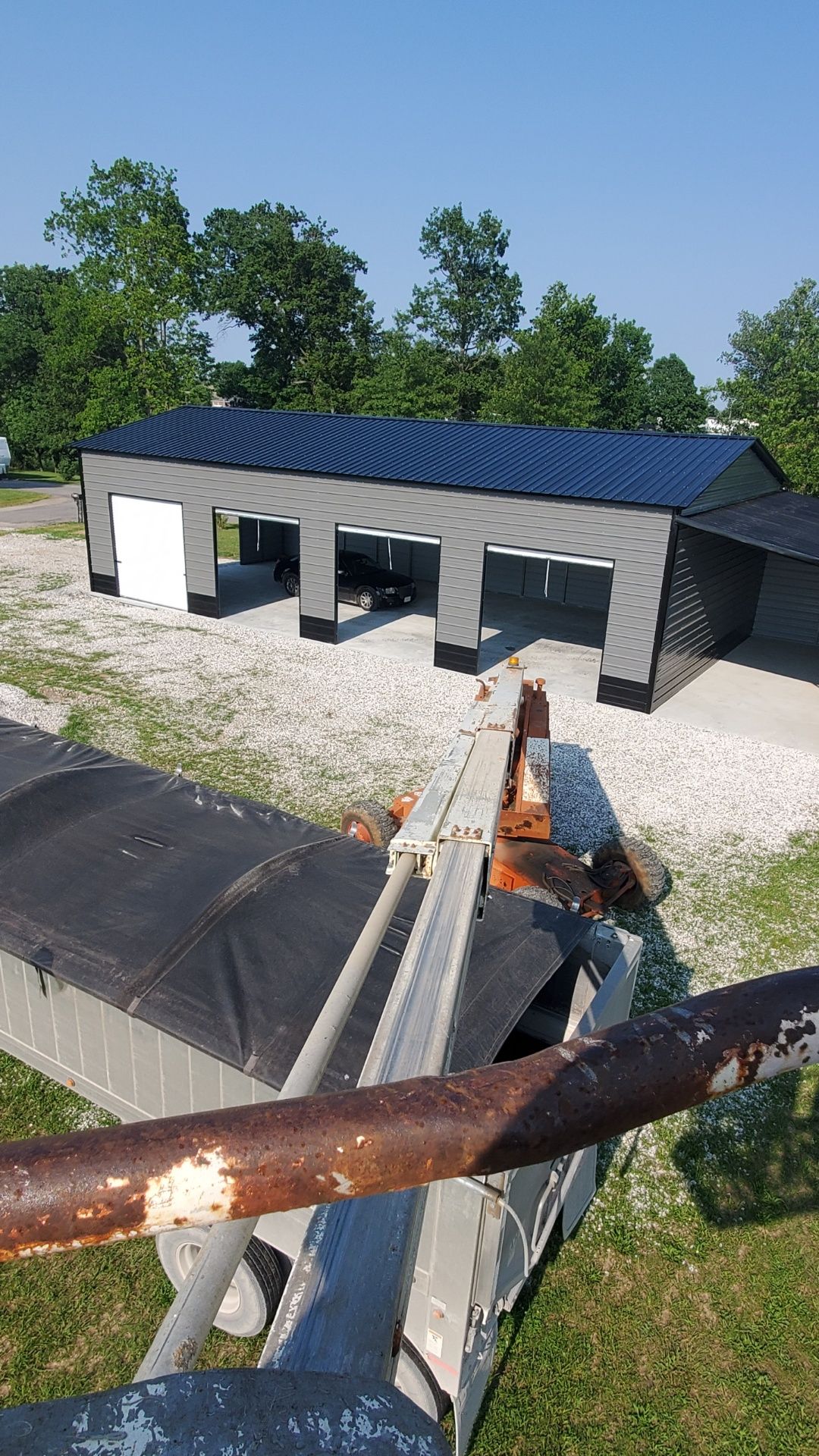 Blue-roofed garage with open bays, seen from a rusty metal structure. Gravel lot, trees in background.