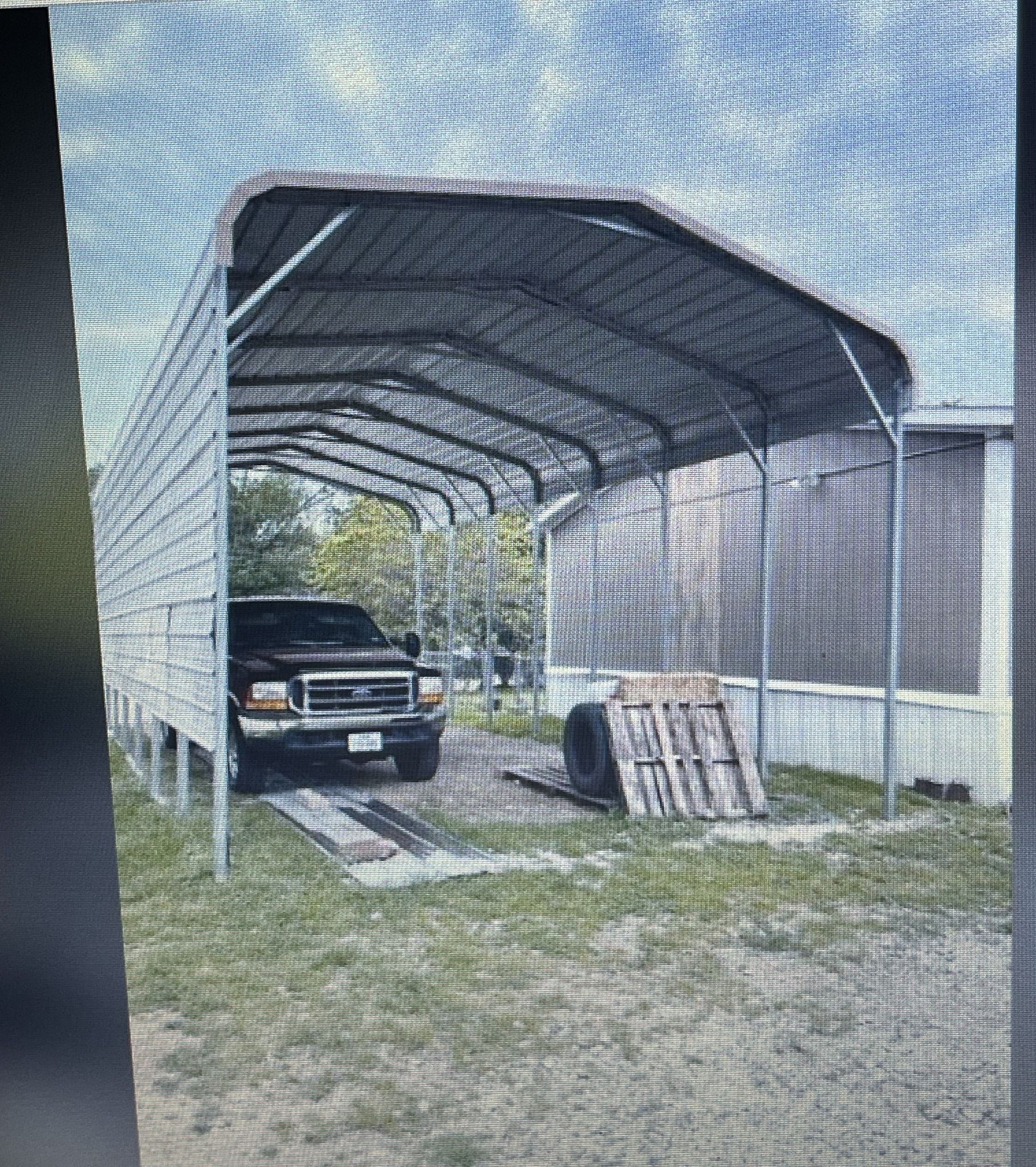 Black truck under a metal carport in a grassy yard next to a white trailer.
